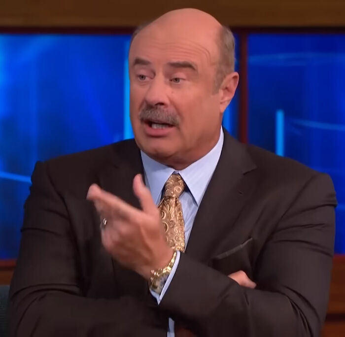 Bald man with mustache in dark suit and patterned tie speaking animatedly on a talk show set.