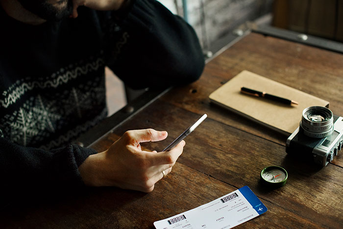 Passenger using a phone at a wooden table with tickets, notebook, camera, and compass nearby. Passenger using a phone at a wooden table with tickets, notebook, camera, and compass nearby.