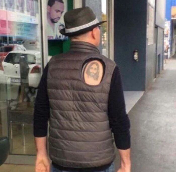 Man in a hat with a hole in his vest revealing a tattoo, showcasing an epic tattoo fail on a city street.