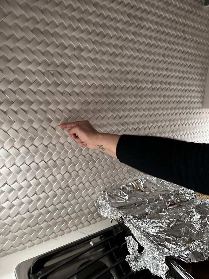 Hand pointing at textured kitchen backsplash with narrow gaps that are difficult to clean near stove covered in foil.