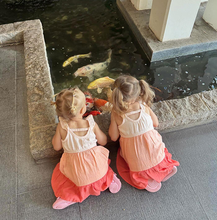 Two young children, connected to Bobbi Althoff&rsquo;s husband, sitting by a koi pond, wearing matching dresses, gazing at colorful fish.