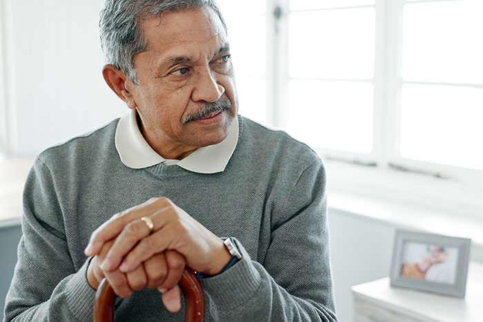Elderly man in a thoughtful pose, sitting indoors, holding a cane with a framed photo in the background.