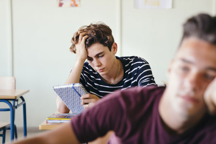Picked-on teen in classroom silently plotting, notebook in hand, bully unaware in foreground.