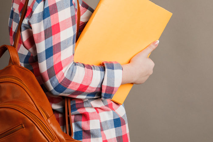 Teen holding yellow folder, wearing a plaid shirt and backpack, symbolizes an academic setting involving a picked-on student.