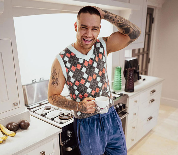 Man with tattoos in kitchen, holding a mug, smiling, and wearing a patterned vest and blue pants. Man with tattoos in kitchen, holding a mug, smiling, and wearing a patterned vest and blue pants.