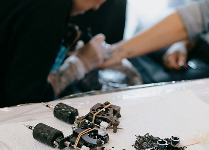 Tattoo artist working on a woman&rsquo;s arm with tattoo equipment on table.