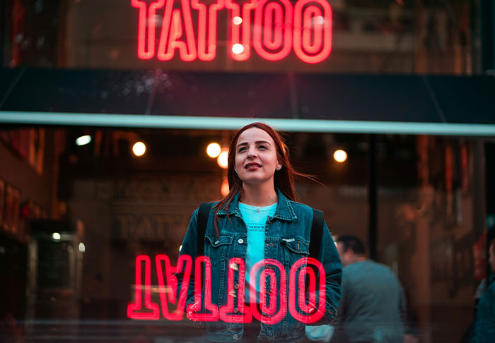 Woman standing outside tattoo shop with bright neon sign reflection.