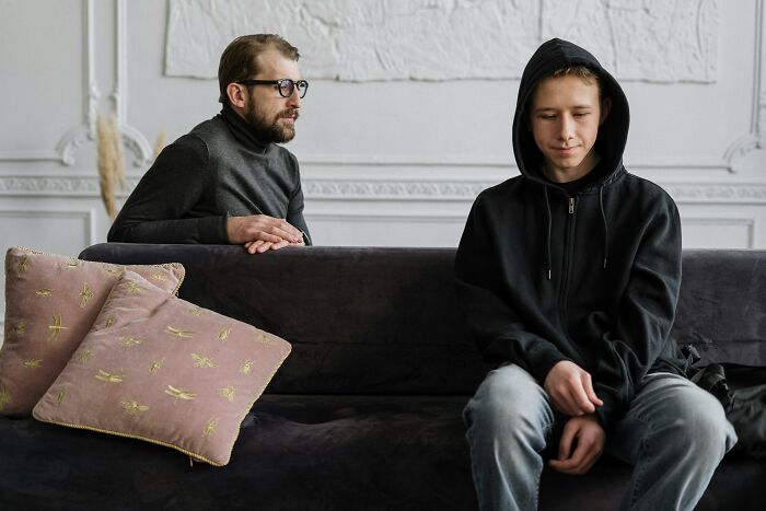 Man and boy in a quiet room, the boy sits on a couch wearing a black hoodie, reflecting on autism's impact on family dynamics. Man and boy in a quiet room, the boy sits on a couch wearing a black hoodie, reflecting on autism's impact on family dynamics.
