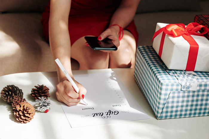 Woman creating a Christmas anti-list, surrounded by gift boxes, holding a phone and pen.