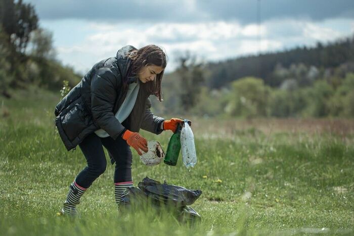 A woman picking up litter in a grass field, wearing gloves and a jacket; an example of things people might dislike.