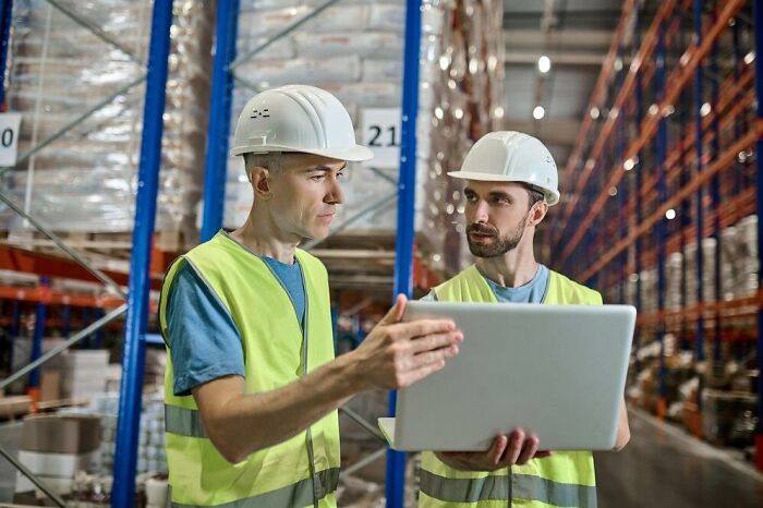 Two workers in a warehouse, wearing safety gear, discussing something on a laptop.