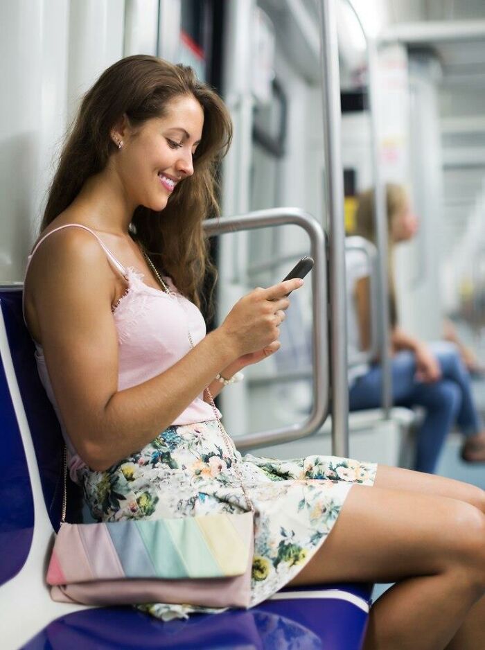 Young woman smiling, using phone on subway, floral skirt, pastel bag; things that make people frustrated in public spaces.