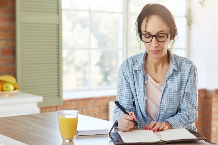 Woman in glasses writing in a notebook at a kitchen table, representing risky decision planning.
