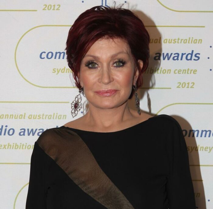 A celebrity at an awards event, wearing a black dress with a brown accent and statement earrings.