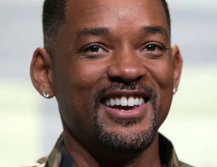 Smiling celebrity at a public event, wearing earrings and a camouflage shirt.