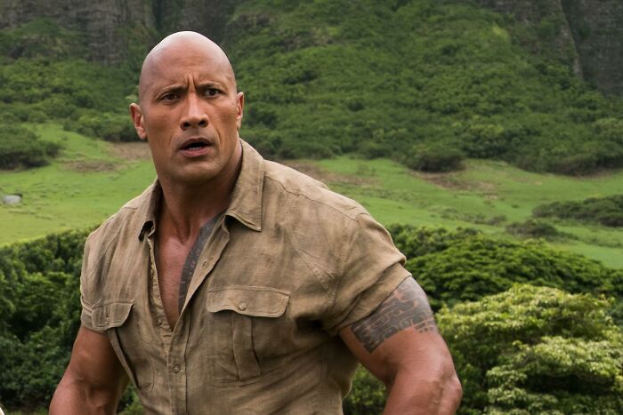 Bald man in rugged shirt looks focused, set against a lush green landscape.