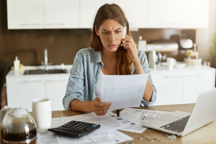 Woman in a kitchen, holding a paper, looking concerned, surrounded by bills and a laptop, represents societal double standards.
