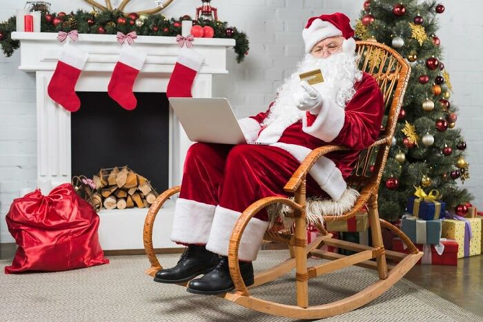 Santa Claus in a rocking chair with a laptop and credit card, in front of a fireplace with stockings, showcasing double standards.