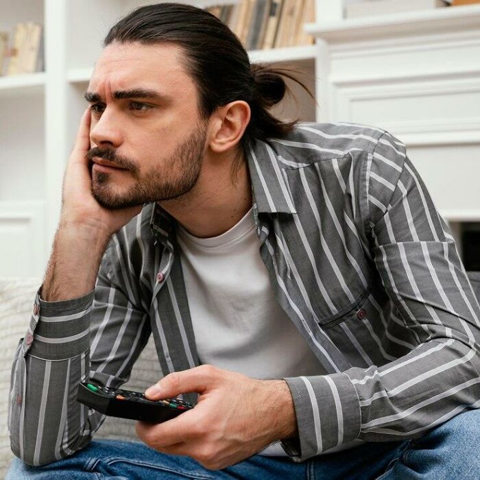 Man in a striped shirt, holding a remote, pondering double standards in society while sitting on a couch.