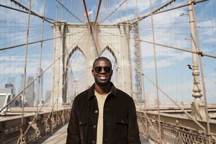 Man on the Brooklyn Bridge, wearing sunglasses and a brown jacket, symbolizing societal double standards.