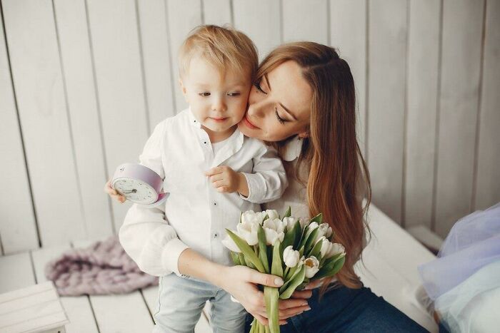 Woman holding a bouquet of tulips, hugging a child with an alarm clock, illustrating double standards in society.