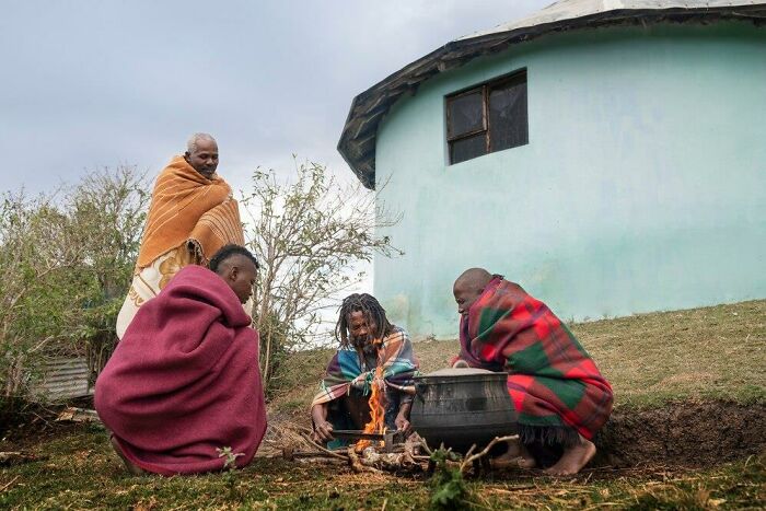 People wrapped in colorful blankets sitting near a fire, discussing iron levels and veganism near a round hut.
