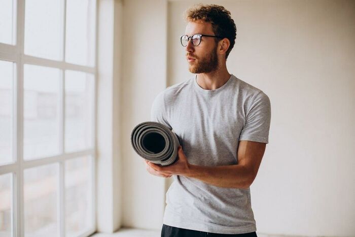 Man in a gray shirt holding a yoga mat near a window, contemplating low iron levels and veganism.