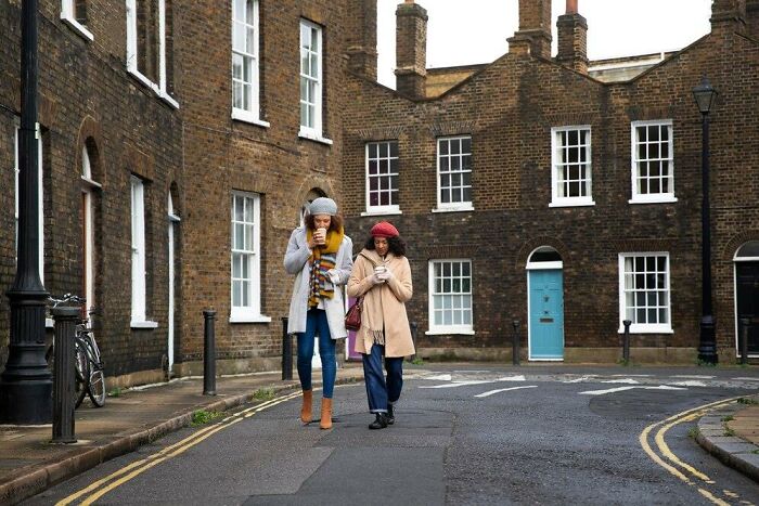 Two women walking in a quiet British street, each engrossed in their phones, surrounded by brick buildings and a parked bike.