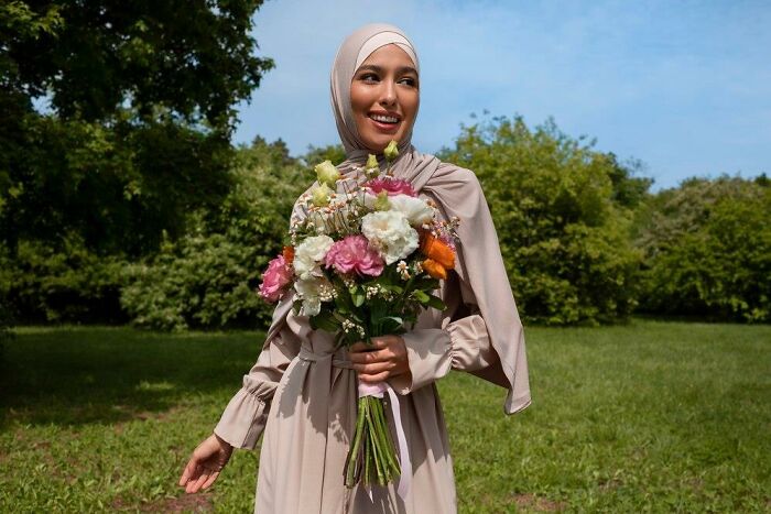 Woman in a beige dress holding a colorful bouquet, standing in a lush green park, smiling under a clear blue sky.
