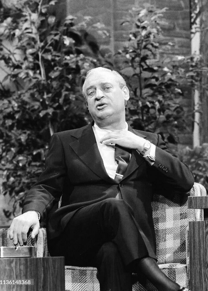 A man in a suit sits on a talk show set, gesturing expressively, sharing stories about his experiences with celebrities.