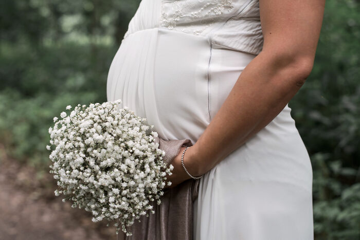 Pregnant bridesmaid in white dress holds bouquet at a wedding.
