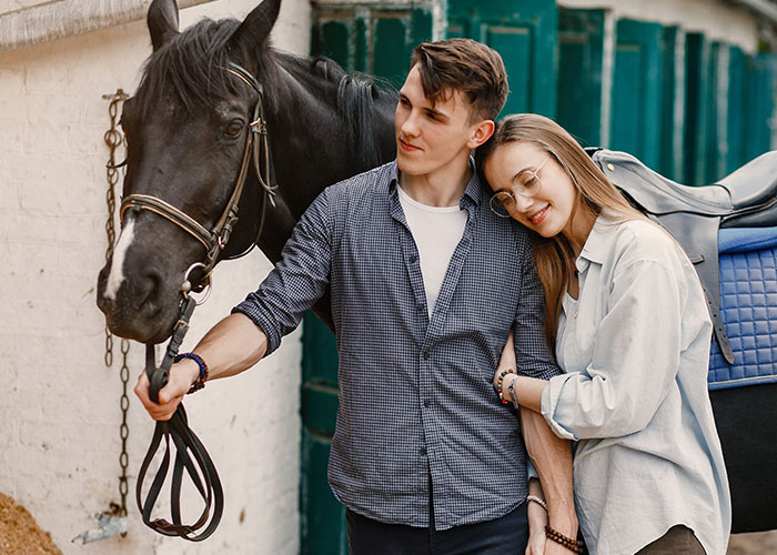 A couple standing next to a black horse in a stable, portraying a relationship dynamic as part of a wild cheating story.