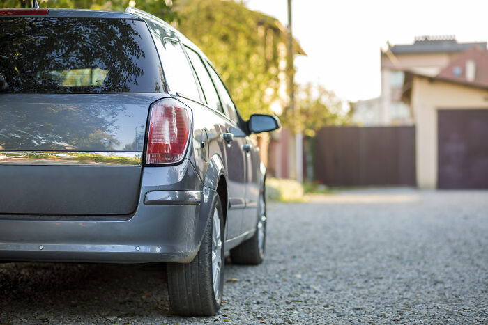 Grey car parked on a gravel driveway near houses, with trees in the background.