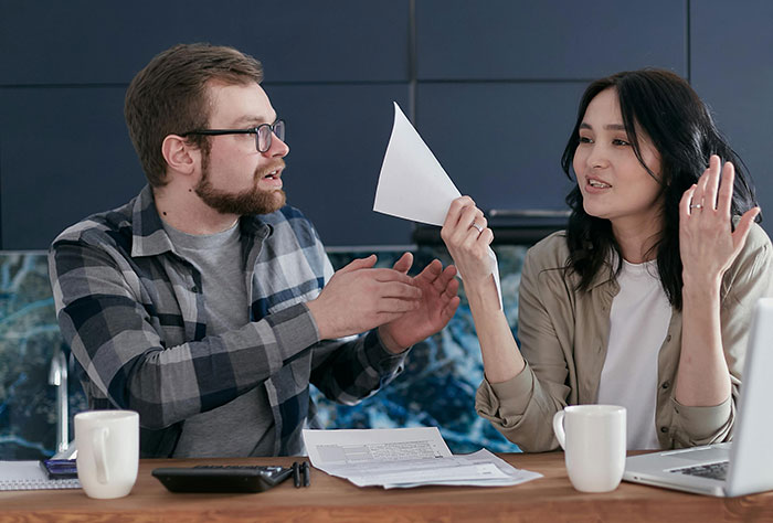 Couple discussing real estate paperwork at a table with coffee mugs and a laptop, highlighting home ownership conflict.