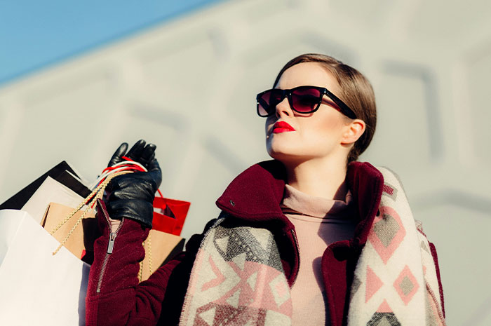 Woman in sunglasses, holding shopping bags, wearing a patterned scarf and burgundy coat, representing independence.