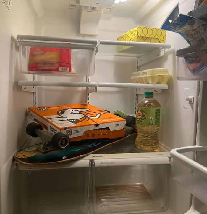 Inside a chaotic Dude Fridge: pizza box on skateboard, oil bottle, and eggs stacked haphazardly.