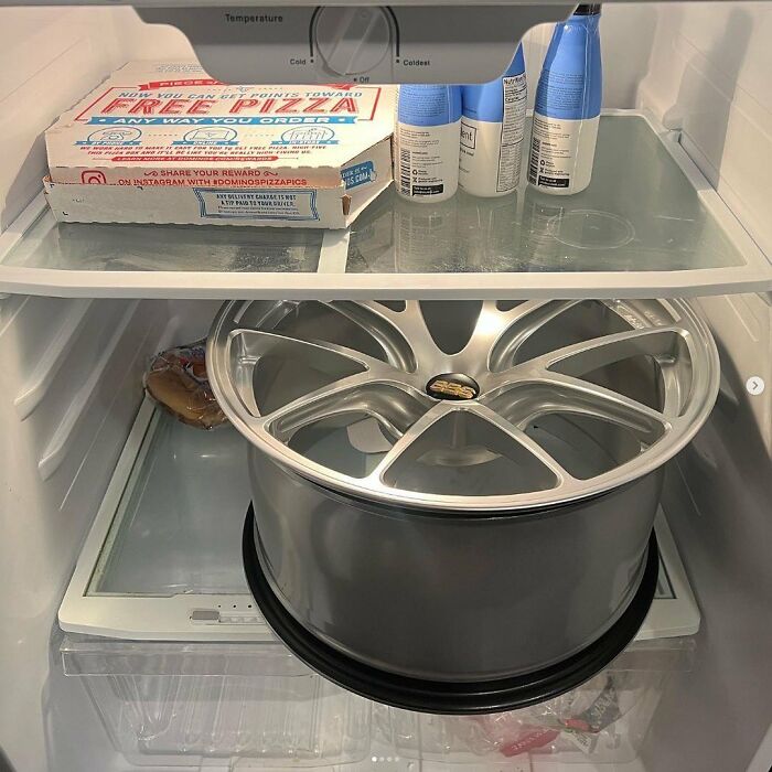 Chaotic fridge with a pizza box, car wheel rim, and hygiene products, reflecting the 'Dude Fridges' theme.