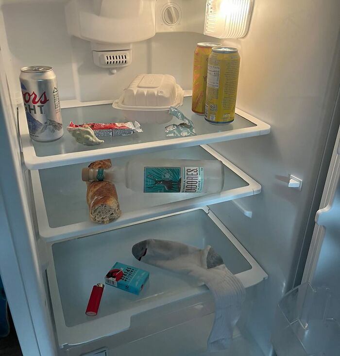 Chaotic dude fridge with beer, bread, vodka, lighter, and a single sock on near-empty shelves.