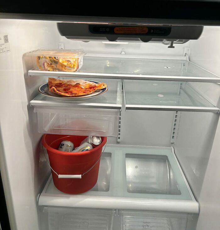 Inside a chaotic 'Dude Fridge': sparse contents include pizza, a takeout container, and a red bucket holding cans.