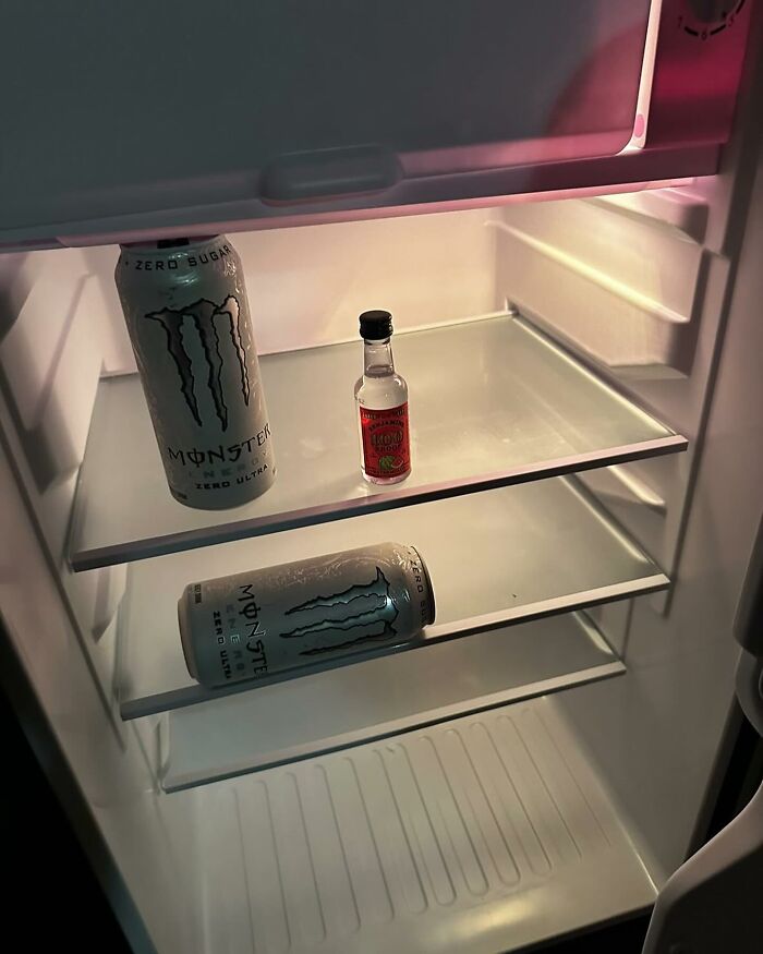 Inside a dude fridge: two energy drinks and a mini bottle of alcohol on empty shelves.