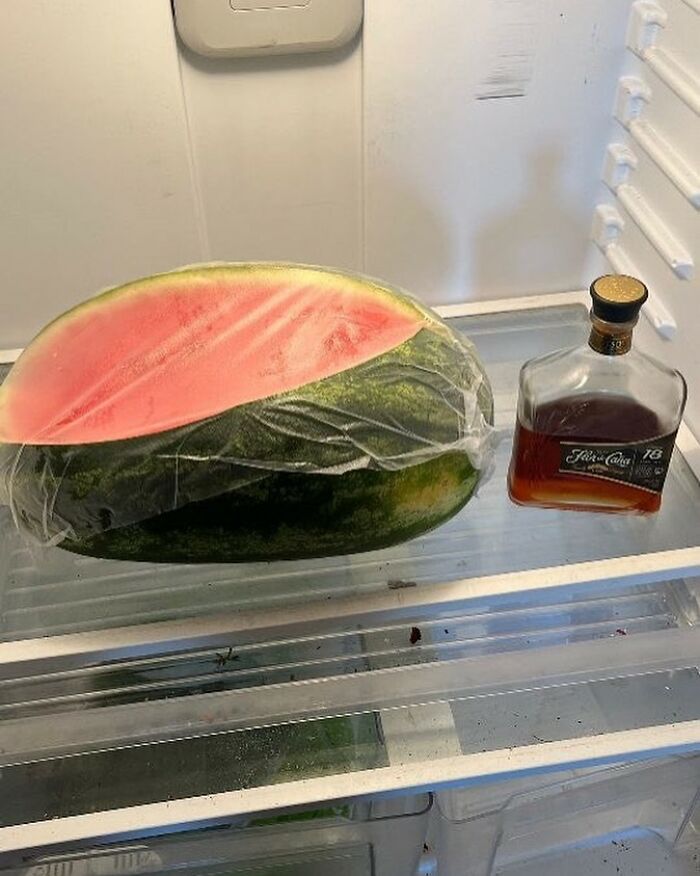 Half a watermelon and a bottle of whiskey in an open refrigerator, exemplifying a 'dude fridge'.
