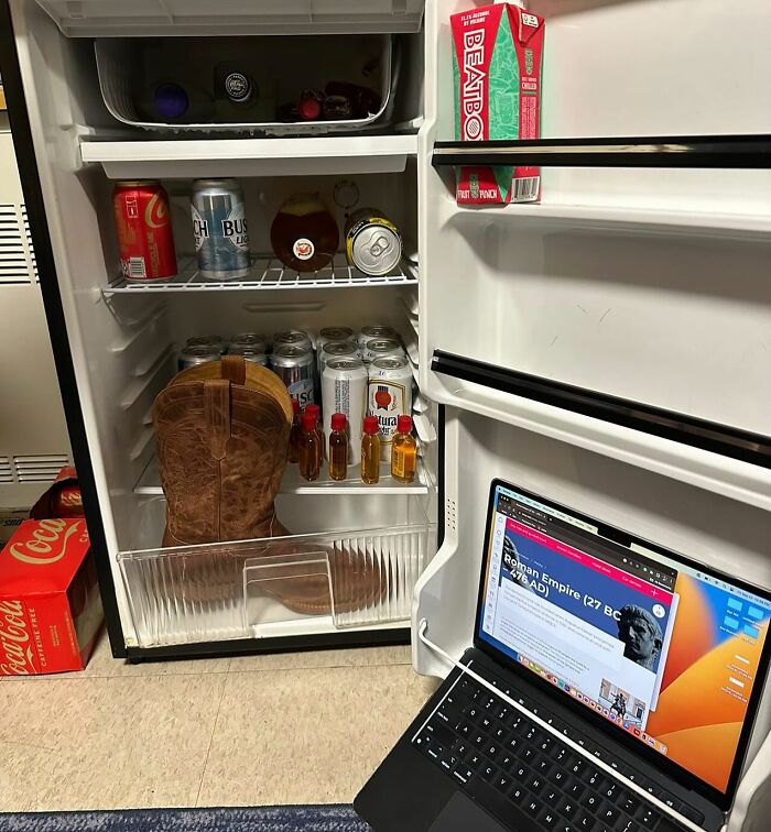 Chaotic dude fridge with drinks, a boot, and a laptop showing a Roman Empire article.