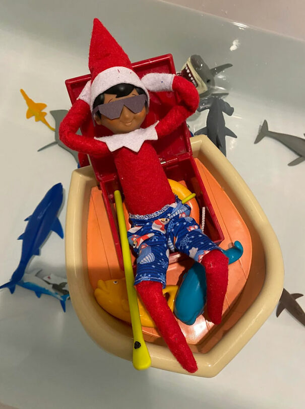 Elf on the Shelf relaxing in a toy boat with sunglasses and swim trunks surrounded by toy sharks in a bathtub.