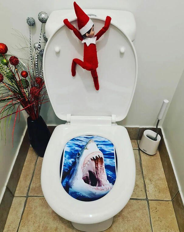 Elf On The Shelf clings to toilet seat with shark mouth decal creating a funny mischief scene in bathroom.