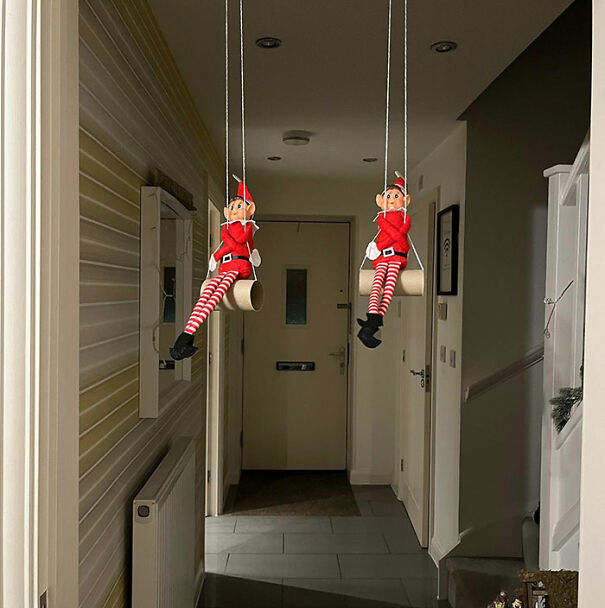 Two Elf On The Shelf dolls sitting on swings made from toilet paper rolls hanging from the ceiling in a hallway.