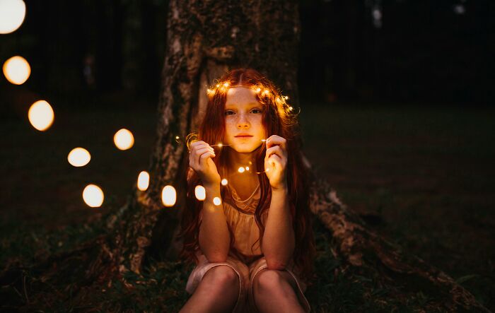 Girl with fairy lights sitting by a tree at dusk, evoking smell-happiness ambiance.