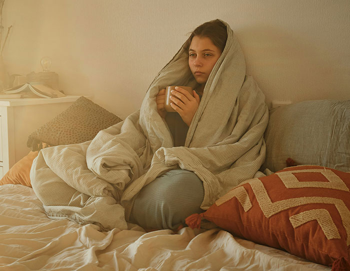 Sad woman wrapped in a blanket, holding a mug, reflecting on friendship issues in her bedroom.