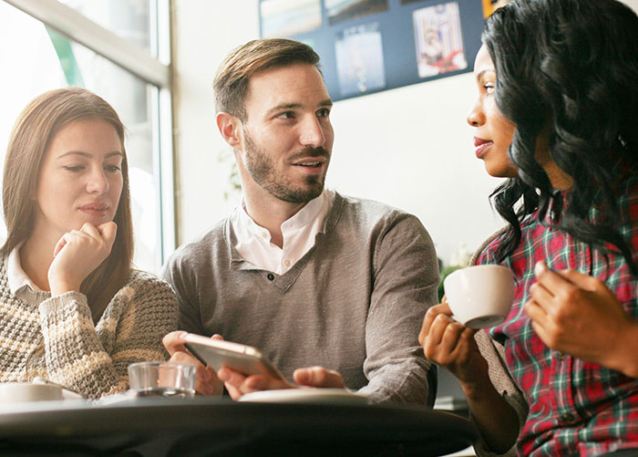 Three friends in a cafe; one holds a phone, sharing an accidental text revealing friendship issues.