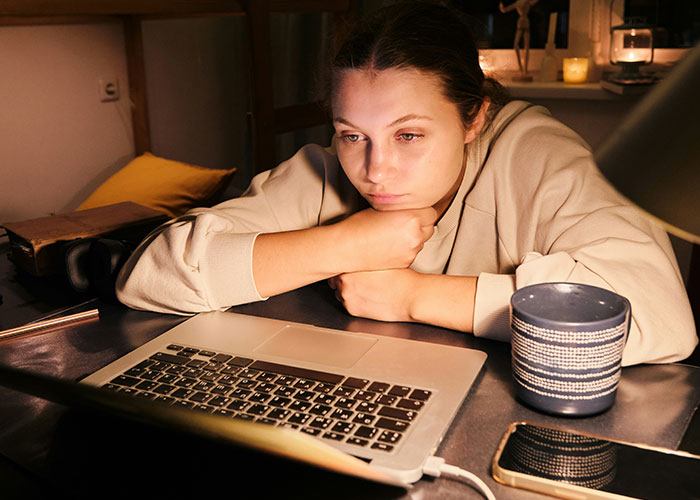 Woman watching TikTok trend video on laptop, looking stunned and thoughtful in a dimly lit room.