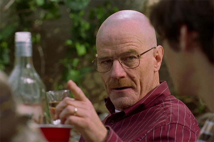 Bald actor in glasses, holding a drink, wearing a red striped shirt, flawlessly embodying a character in a garden setting.